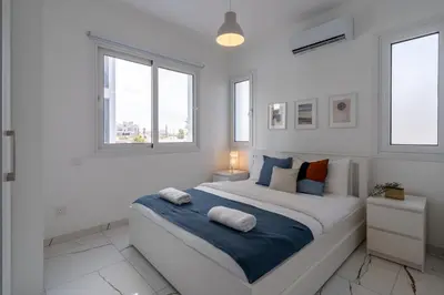 a white bedroom with a large bed with two windows at Artemis 1-Bedroom Apartment in Larnaca in Larnaka