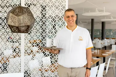 a man in a white shirt holding a tray at SUNWING Sandy Bay Beach Village in Ayia Napa