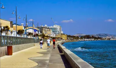 two people walking down a sidewalk next to the water at ONAR mackenzie 1 in Larnaka
