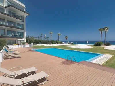 a swimming pool next to a building with chairs and the ocean at Apartment Luxuriöse Wohnung am Strand von Thalassa by Interhome in Protaras