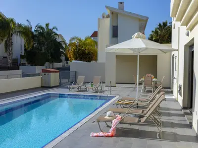 a swimming pool with chairs and an umbrella next to a house at Protaras Villa Theodora 1 in Protaras